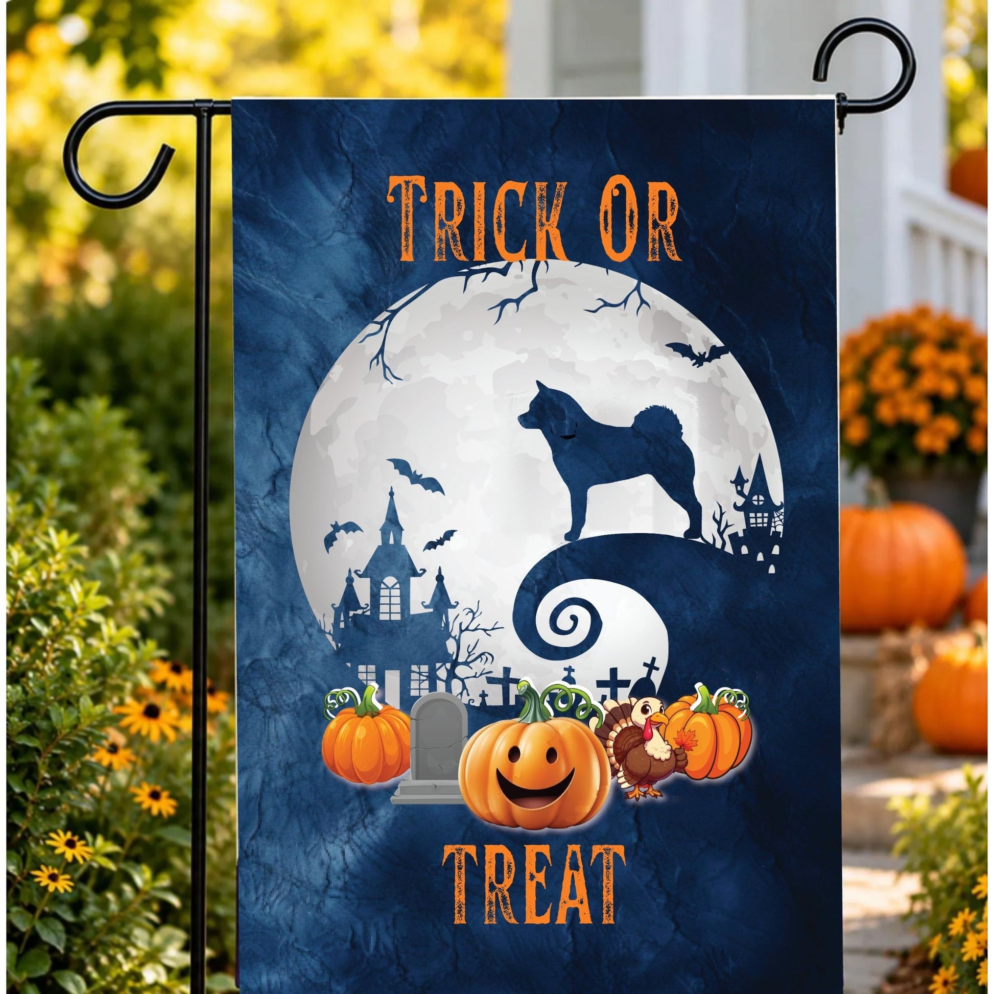 A vertical garden flag with a Halloween design, featuring a dark blue background, a large white full moon, a akita silhouette, haunted houses, bats, pumpkins, and the words 'TRICK OR TREAT'. The flag is displayed on a black metal garden flag stand outdoors in a yard setting with pumpkins and flowers.