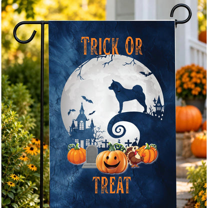 A vertical garden flag with a Halloween design, featuring a dark blue background, a large white full moon, a akita silhouette, haunted houses, bats, pumpkins, and the words 'TRICK OR TREAT'. The flag is displayed on a black metal garden flag stand outdoors in a yard setting with pumpkins and flowers.